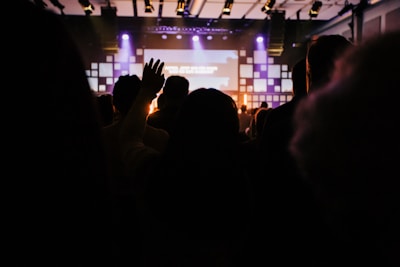 A dimly lit indoor venue with a crowd of people facing a stage. Warm lighting and a large screen are visible in the background. One person has their hand raised, suggesting engagement or participation.