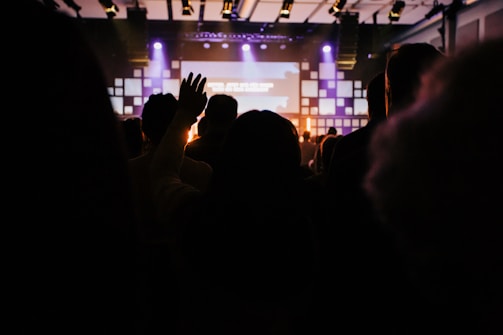 A dimly lit indoor venue with a crowd of people facing a stage. Warm lighting and a large screen are visible in the background. One person has their hand raised, suggesting engagement or participation.