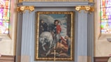 A framed painting set within an ornate and decorative architectural setting. The painting depicts a person on horseback distributing a red cloak to a kneeling figure. The background features an atmospheric sky and other figures watching the scene. Rich stained glass windows flank the painting and add vibrant colors to the surrounding area.