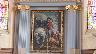 A framed painting set within an ornate and decorative architectural setting. The painting depicts a person on horseback distributing a red cloak to a kneeling figure. The background features an atmospheric sky and other figures watching the scene. Rich stained glass windows flank the painting and add vibrant colors to the surrounding area.