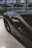 A sleek, dark-colored sports car is parked indoors on a polished marble floor. The car features aerodynamic curves and a prominent air intake on the side. Through the large window, several other vehicles can be seen parked outside on a road, alongside a commercial building.