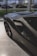A sleek, dark-colored sports car is parked indoors on a polished marble floor. The car features aerodynamic curves and a prominent air intake on the side. Through the large window, several other vehicles can be seen parked outside on a road, alongside a commercial building.