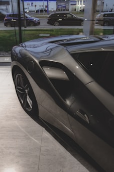 A sleek, dark-colored sports car is parked indoors on a polished marble floor. The car features aerodynamic curves and a prominent air intake on the side. Through the large window, several other vehicles can be seen parked outside on a road, alongside a commercial building.