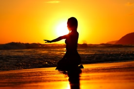 A silhouette of a person performing a yoga pose on the beach at sunset. The vibrant orange and yellow hues of the sunset create a dramatic backdrop, with the ocean waves gently rolling onto the shore.