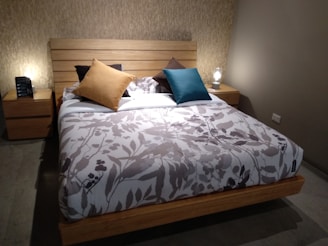 A cozy bedroom featuring a wooden bed with a floral patterned bedspread. The bed is adorned with several pillows in different colors including mustard yellow, teal, and brown. It is flanked by two wooden nightstands, each with a small lamp emitting warm light. The walls are painted in a neutral tone, enhancing the room's warm and inviting atmosphere.