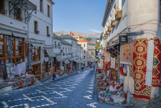 A cobblestone street lined with traditional shops displaying colorful textiles, carpets, and souvenirs. The architecture of the buildings is historic, with wooden window frames and white facades. There are a few people casually walking along the street, and the overall setting suggests a vibrant market atmosphere.
