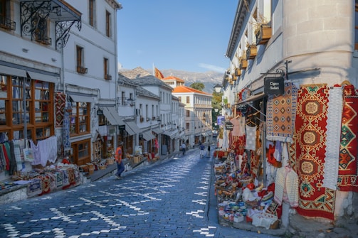 A cobblestone street lined with traditional shops displaying colorful textiles, carpets, and souvenirs. The architecture of the buildings is historic, with wooden window frames and white facades. There are a few people casually walking along the street, and the overall setting suggests a vibrant market atmosphere.
