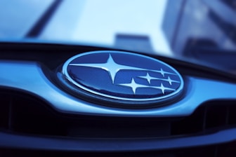 A close-up of a car emblem featuring several silver stars arranged on a blue background with a metallic border. The background appears slightly reflective, and the image has a cool blue tone.