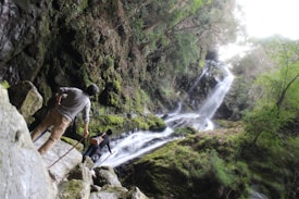 A lush, verdant landscape featuring a cascading waterfall surrounded by dense green foliage. Two hikers are seen navigating rocky terrain, with one person using a walking stick, as they make their way towards the waterfall.