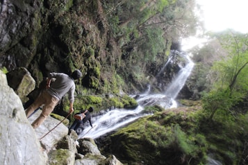A lush, verdant landscape featuring a cascading waterfall surrounded by dense green foliage. Two hikers are seen navigating rocky terrain, with one person using a walking stick, as they make their way towards the waterfall.