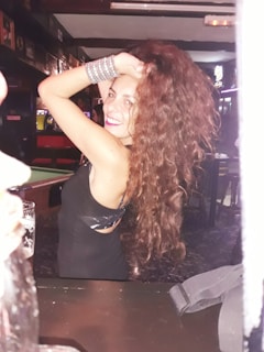 A person with long curly hair, wearing a black dress and silver bracelets, smiling and posing with one arm raised in a lively bar setting.