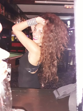 A person with long curly hair, wearing a black dress and silver bracelets, smiling and posing with one arm raised in a lively bar setting.