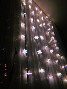 A string of decorative snowflake-shaped lights hanging vertically against a sheer curtain. The warm glow of the lights contrasts with the dark background, creating a cozy and festive atmosphere. The lights are evenly spaced, casting faint shadows and reflections on the curtain.