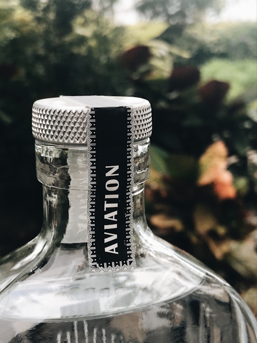 A close-up of a bottle with a metallic cap featuring a textured design. A label with the word 'Aviation' is affixed to the neck of the bottle. The background is blurred, consisting of greenery and earthy tones.