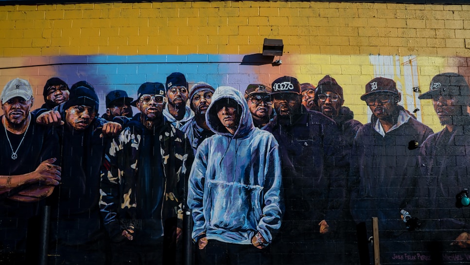 A mural depicting a group of people wearing hooded sweatshirts, jackets, and hats. The group stands closely together against a painted brick wall background, creating a sense of unity. The mural features detailed expressions and various clothing styles, showcasing realistic and vibrant artistry.