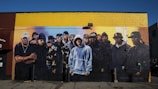 A mural painted on a building wall features a group of individuals wearing various urban and hip-hop style clothing, such as caps, hoodies, and jackets. The mural displays a diverse blend of expressions, from serious to neutral, and encompasses a broad spectrum of styles. The wall itself is painted in sections of yellow and brown, providing a vibrant backdrop for the detailed, life-sized figures depicted in darker colors.