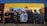 A mural painted on a building wall features a group of individuals wearing various urban and hip-hop style clothing, such as caps, hoodies, and jackets. The mural displays a diverse blend of expressions, from serious to neutral, and encompasses a broad spectrum of styles. The wall itself is painted in sections of yellow and brown, providing a vibrant backdrop for the detailed, life-sized figures depicted in darker colors.