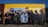 A mural painted on a building wall features a group of individuals wearing various urban and hip-hop style clothing, such as caps, hoodies, and jackets. The mural displays a diverse blend of expressions, from serious to neutral, and encompasses a broad spectrum of styles. The wall itself is painted in sections of yellow and brown, providing a vibrant backdrop for the detailed, life-sized figures depicted in darker colors.