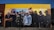 A mural painted on a building wall features a group of individuals wearing various urban and hip-hop style clothing, such as caps, hoodies, and jackets. The mural displays a diverse blend of expressions, from serious to neutral, and encompasses a broad spectrum of styles. The wall itself is painted in sections of yellow and brown, providing a vibrant backdrop for the detailed, life-sized figures depicted in darker colors.