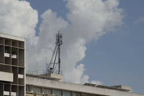 A concrete building with rectangular balconies extends into the sky under large, fluffy clouds. Atop the building, there is a tall communication tower with various antennas and satellite dishes.