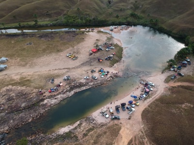 Aerial view of a riverbend surrounded by grassy hills and scattered vehicles, with tents set up along the riverbanks. People are gathered near the water, engaging in recreational activities. The landscape is a mix of green vegetation and dry areas.