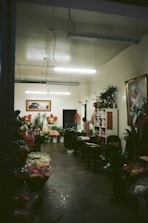 A dimly lit flower shop interior with a variety of colorful flowers displayed in buckets and on shelves. The room has a simple decor with fluorescent lights on the ceiling, and artwork hanging on the walls. A person is standing near a shelf organizing flowers or plants.