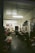 A dimly lit flower shop interior with a variety of colorful flowers displayed in buckets and on shelves. The room has a simple decor with fluorescent lights on the ceiling, and artwork hanging on the walls. A person is standing near a shelf organizing flowers or plants.