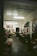 A dimly lit flower shop interior with a variety of colorful flowers displayed in buckets and on shelves. The room has a simple decor with fluorescent lights on the ceiling, and artwork hanging on the walls. A person is standing near a shelf organizing flowers or plants.