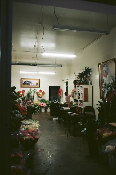 A dimly lit flower shop interior with a variety of colorful flowers displayed in buckets and on shelves. The room has a simple decor with fluorescent lights on the ceiling, and artwork hanging on the walls. A person is standing near a shelf organizing flowers or plants.