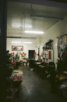 A dimly lit flower shop interior with a variety of colorful flowers displayed in buckets and on shelves. The room has a simple decor with fluorescent lights on the ceiling, and artwork hanging on the walls. A person is standing near a shelf organizing flowers or plants.