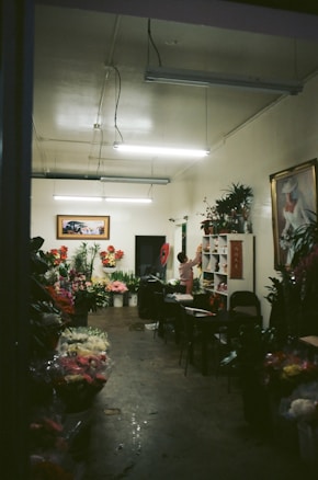 A dimly lit flower shop interior with a variety of colorful flowers displayed in buckets and on shelves. The room has a simple decor with fluorescent lights on the ceiling, and artwork hanging on the walls. A person is standing near a shelf organizing flowers or plants.