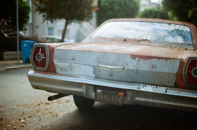 An aged Ford car is parked on a quiet residential street. The car appears weathered and rusted, with visible paint peeling off. There are old California license plates and a bumper sticker that reads 'Oakland Raiders'. The background features trees and residential buildings.