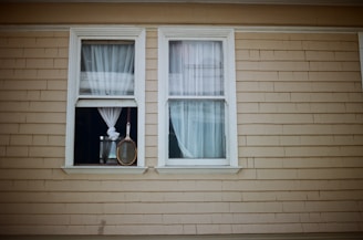 Two windows with white curtains are set in a beige wood-paneled wall. One window is partially open, and a tennis racket hangs in front of it by its handle.