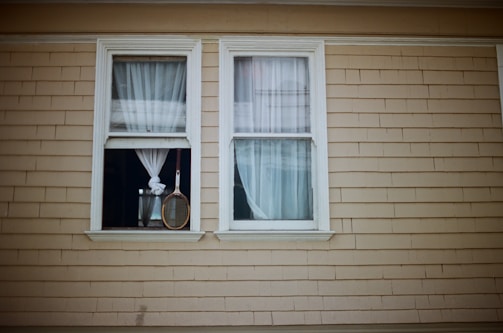 Two windows with white curtains are set in a beige wood-paneled wall. One window is partially open, and a tennis racket hangs in front of it by its handle.