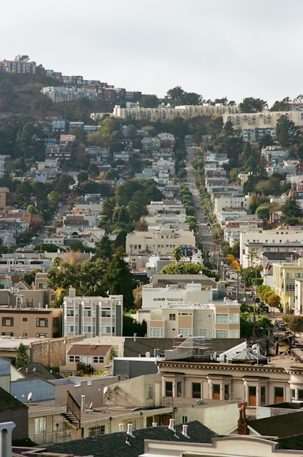 A densely built, urban residential area with rows of apartment buildings descending down a hill. The street is flanked by trees and greenery, and the scene has a variety of architectural styles. The hill in the background is covered with more housing structures, blending into the landscape.