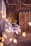 A rustic setting features wooden walls adorned with strings of warm fairy lights. An assortment of pastries and cupcakes, some displayed on tiered trays, are arranged on tables draped with lace and fabric. Floral arrangements and decorative lanterns add to the charming ambiance.