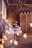 A rustic setting features wooden walls adorned with strings of warm fairy lights. An assortment of pastries and cupcakes, some displayed on tiered trays, are arranged on tables draped with lace and fabric. Floral arrangements and decorative lanterns add to the charming ambiance.
