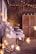 A rustic setting features wooden walls adorned with strings of warm fairy lights. An assortment of pastries and cupcakes, some displayed on tiered trays, are arranged on tables draped with lace and fabric. Floral arrangements and decorative lanterns add to the charming ambiance.
