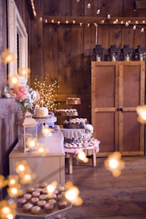 A rustic setting features wooden walls adorned with strings of warm fairy lights. An assortment of pastries and cupcakes, some displayed on tiered trays, are arranged on tables draped with lace and fabric. Floral arrangements and decorative lanterns add to the charming ambiance.