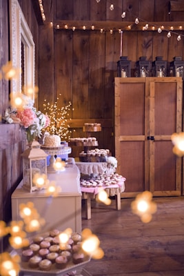 A rustic setting features wooden walls adorned with strings of warm fairy lights. An assortment of pastries and cupcakes, some displayed on tiered trays, are arranged on tables draped with lace and fabric. Floral arrangements and decorative lanterns add to the charming ambiance.