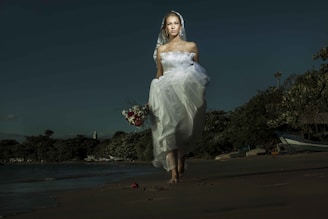 A bride in a white dress and veil is walking barefoot on a beach. She holds a bouquet of red and white flowers, with a serene expression. The background features a shoreline with trees and boats under a dark, moody sky.