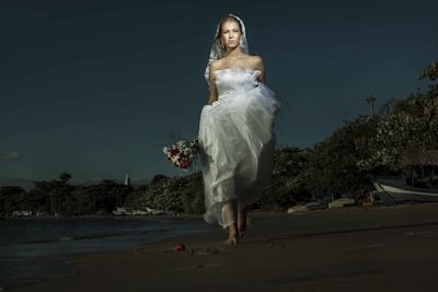 A bride in a white dress and veil is walking barefoot on a beach. She holds a bouquet of red and white flowers, with a serene expression. The background features a shoreline with trees and boats under a dark, moody sky.