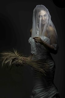 A person dressed in a white wedding gown, partially covered by a delicate, translucent veil. They are holding a bunch of wheat stalks in their hands. The lighting is moody and dramatic, creating shadows that emphasize the texture of the veil and gown.