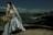 A bride in an elegant white gown and veil is seated on a beach, holding a bouquet of wheat stalks. The background features a city skyline across a body of water, under a cloudy sky. The scene is a juxtaposition of nature and urban landscapes.