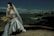 A bride in an elegant white gown and veil is seated on a beach, holding a bouquet of wheat stalks. The background features a city skyline across a body of water, under a cloudy sky. The scene is a juxtaposition of nature and urban landscapes.