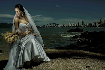 A bride in an elegant white gown and veil is seated on a beach, holding a bouquet of wheat stalks. The background features a city skyline across a body of water, under a cloudy sky. The scene is a juxtaposition of nature and urban landscapes.