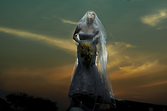 A bride in a detailed lace wedding gown holds a bouquet, veiled and illuminated dramatically against a dusky sky with golden clouds.