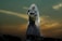 A bride in a detailed lace wedding gown holds a bouquet, veiled and illuminated dramatically against a dusky sky with golden clouds.