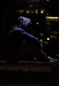 A confident person wearing a dark streetwear hoodie listening to a podcast with city lights in the background at night.