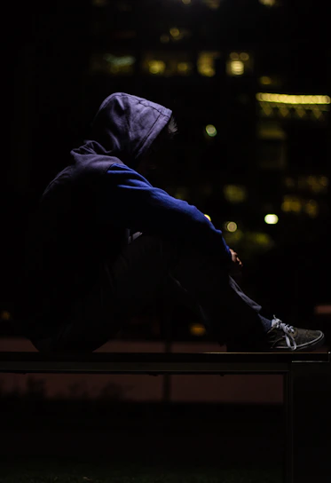 A confident person wearing a dark streetwear hoodie listening to a podcast with city lights in the background at night.